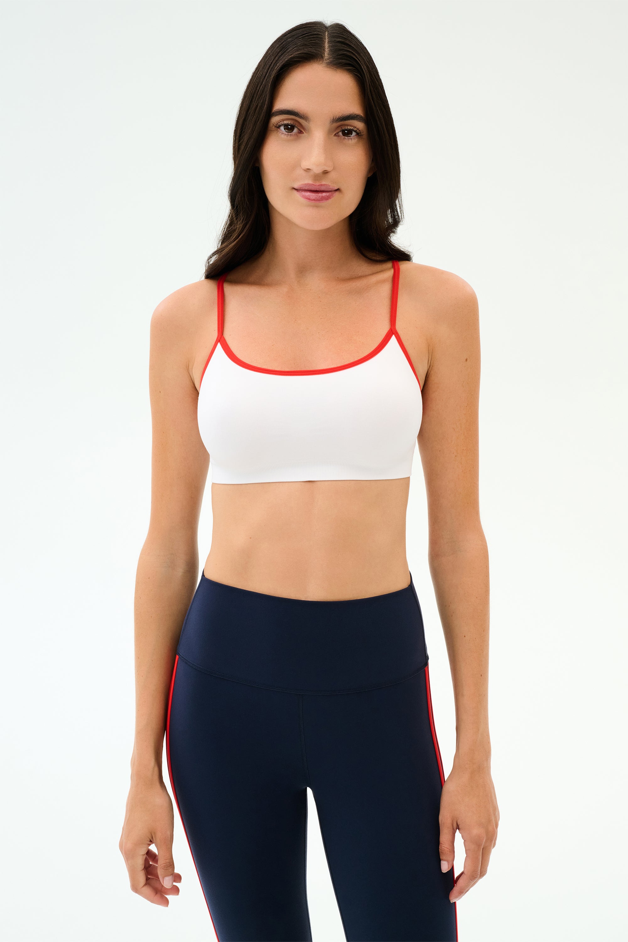 A woman faces forward in a SPLITS59 Loren Seamless Bra - White/Lotus Red, perfect for workouts, paired with high-waisted navy leggings featuring red side stripes, set against a plain light background.