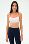 A woman faces forward in a SPLITS59 Loren Seamless Bra - White/Lotus Red, perfect for workouts, paired with high-waisted navy leggings featuring red side stripes, set against a plain light background.