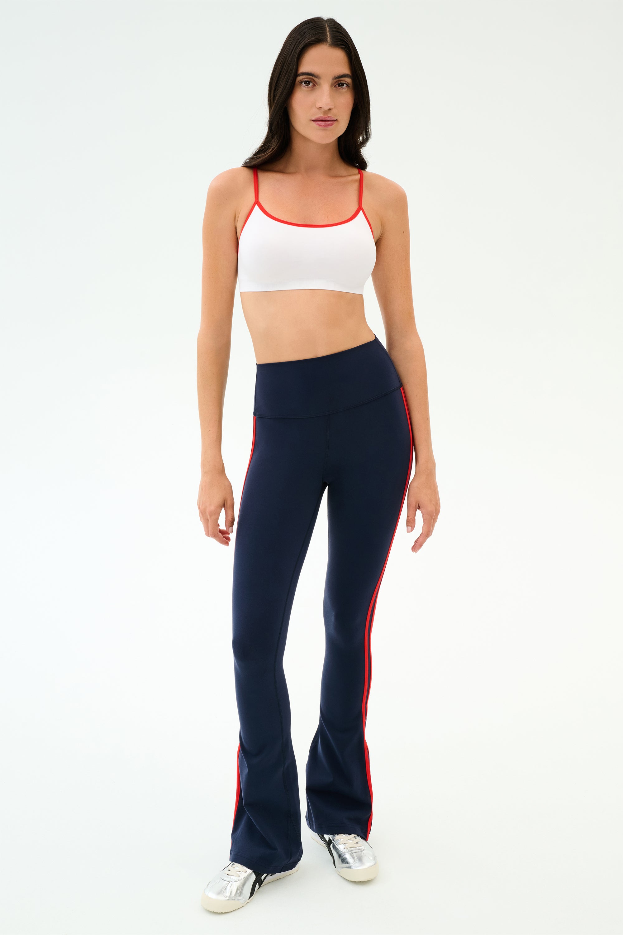 A woman wears the SPLITS59 Loren Seamless Bra in White/Lotus Red, paired with navy blue flare leggings featuring red side stripes and silver sneakers, standing against a plain white background. Perfect for yoga or gym workouts.