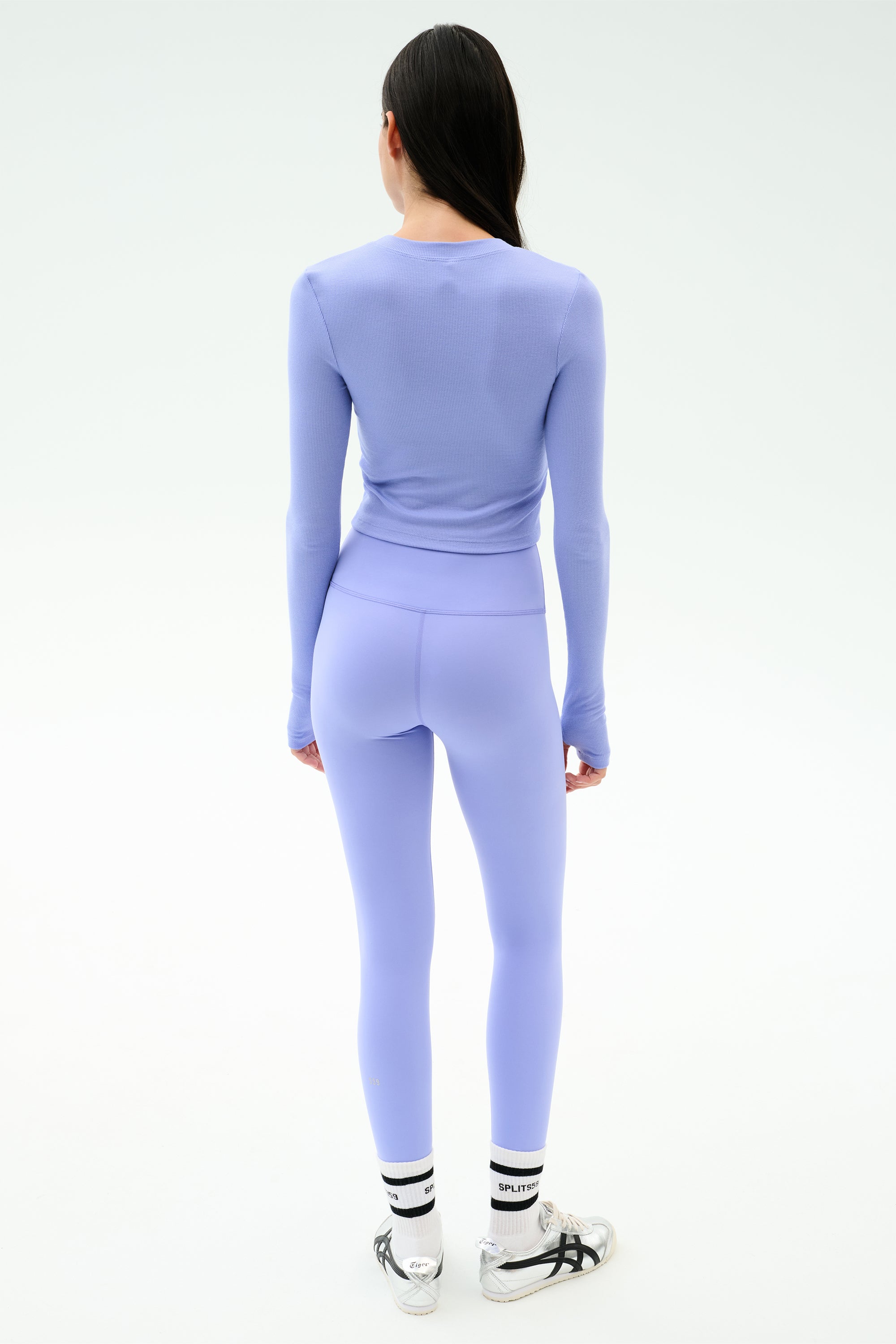 A woman stands facing away, wearing the SPLITS59 Louise Rib Long Sleeve Crop in Purple Haze with matching leggings, white socks with black stripes, and white sneakers against a plain light background.