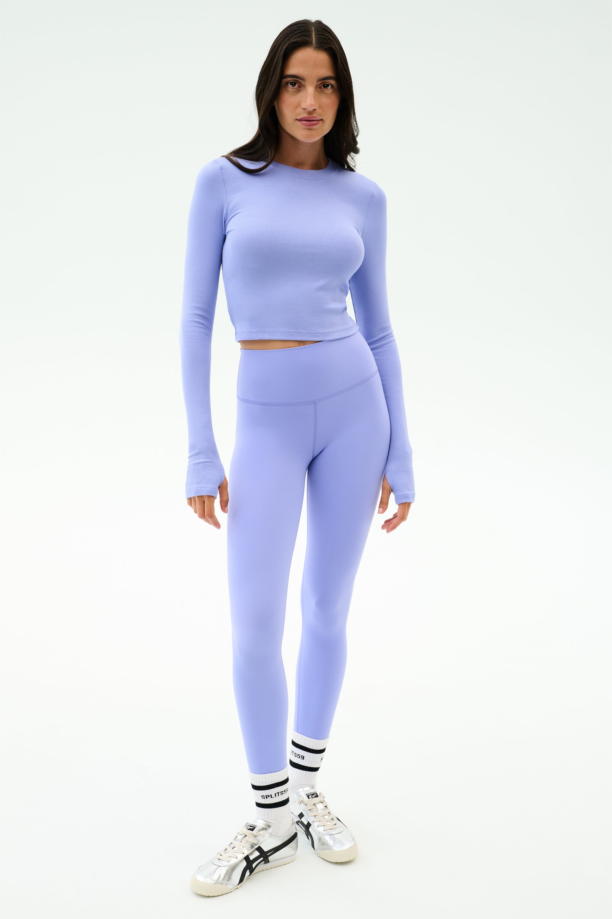 A woman wears the SPLITS59 Louise Rib Long Sleeve Crop in Purple Haze with matching high-waisted leggings, white socks, and silver sneakers against a plain white background.