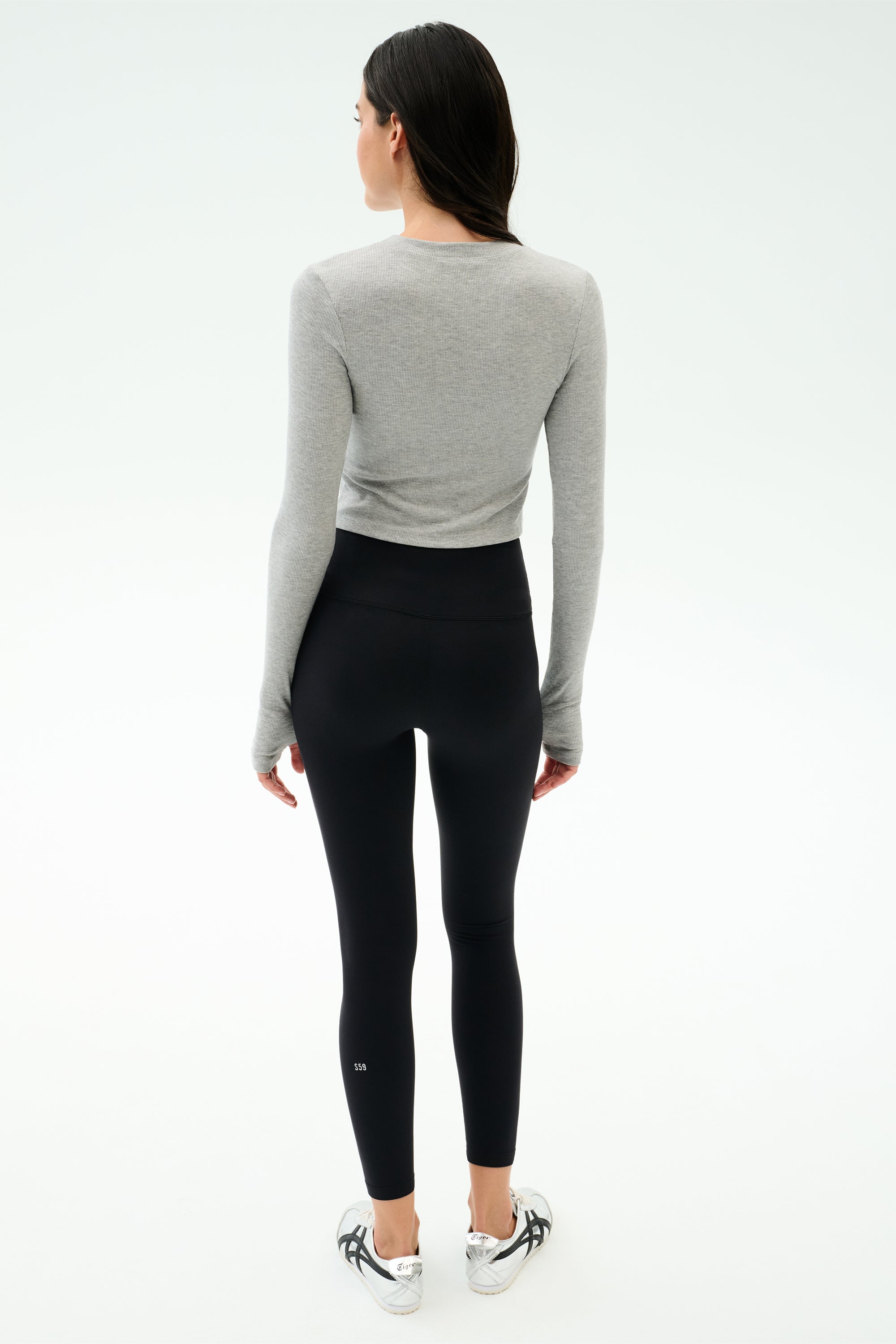 A woman faces away, wearing the SPLITS59 Louise Rib Long Sleeve Crop in Heather Grey, paired with black high-waisted leggings and white athletic shoes, set against a plain light background.