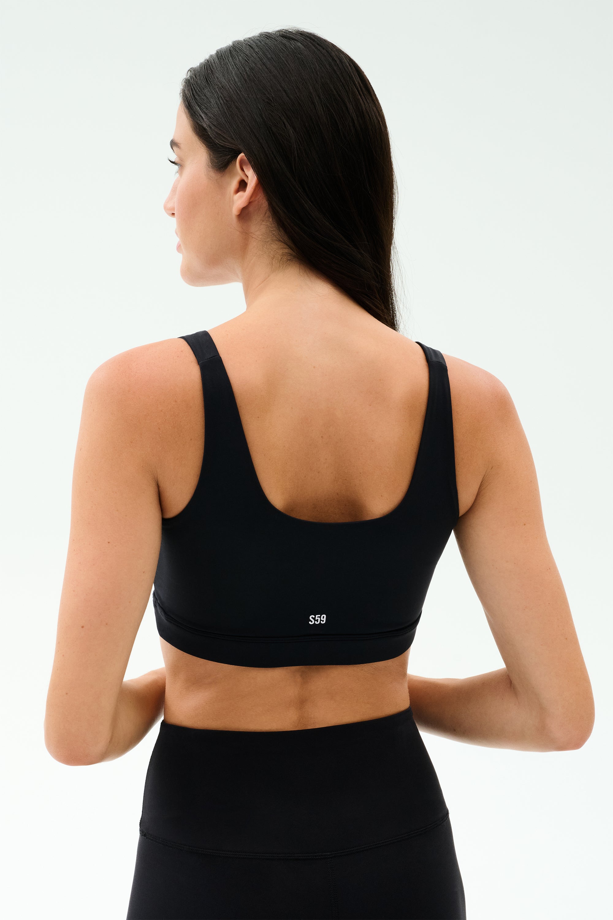A woman with long dark hair, seen from behind, wears the SPLITS59 Sprint Rigor Bra in black and leggings, ready for her gym workout against a plain light background.