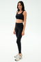 A woman stands against a plain white background wearing the SPLITS59 Sprint Rigor Bra in black, paired with black leggings and white sneakers, ready for her gym workout.
