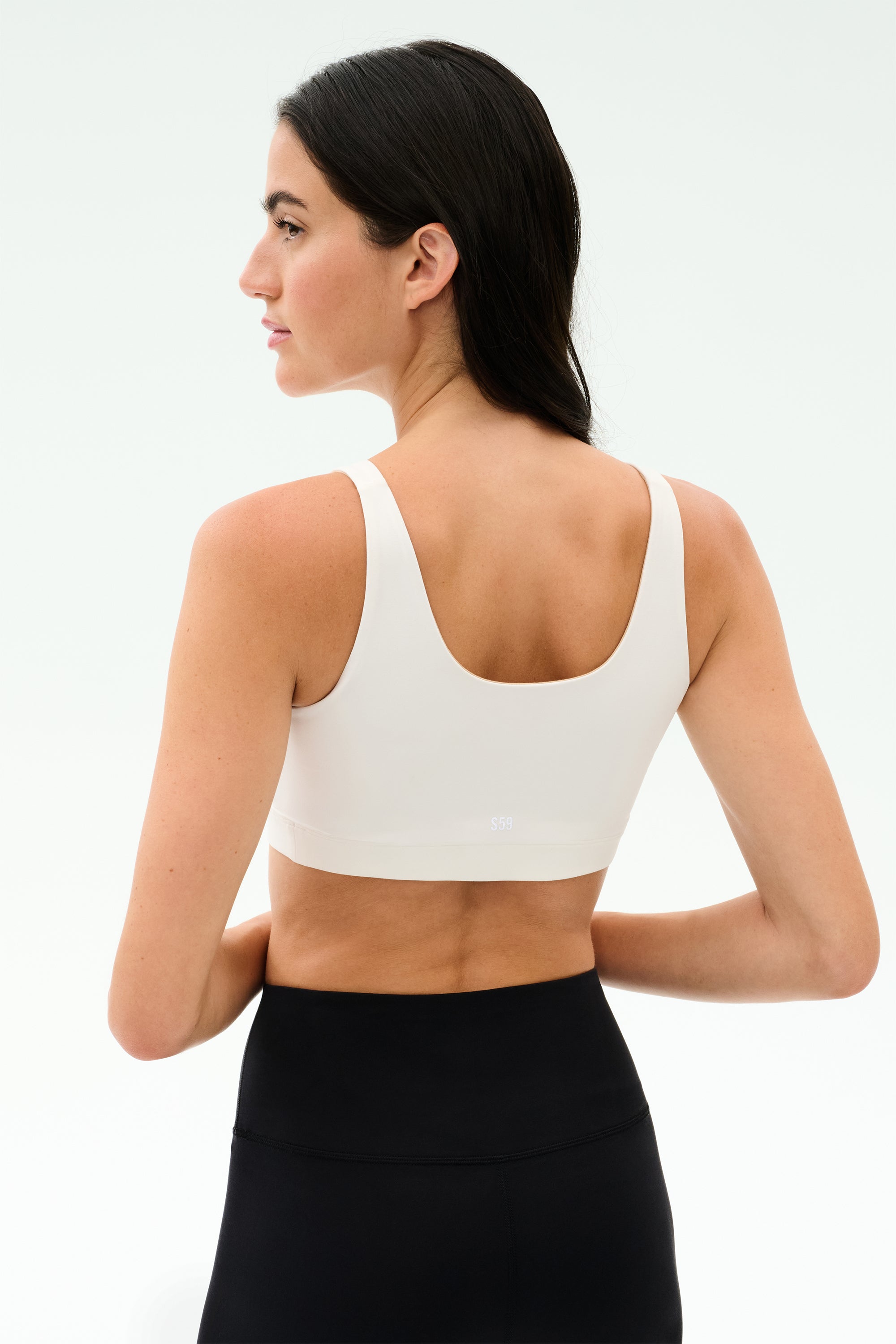 A woman faces away, wearing the SPLITS59 Sprint Rigor Bra in white and black high-waisted leggings against a plain light background—perfect for gym workouts and multisport performance.
