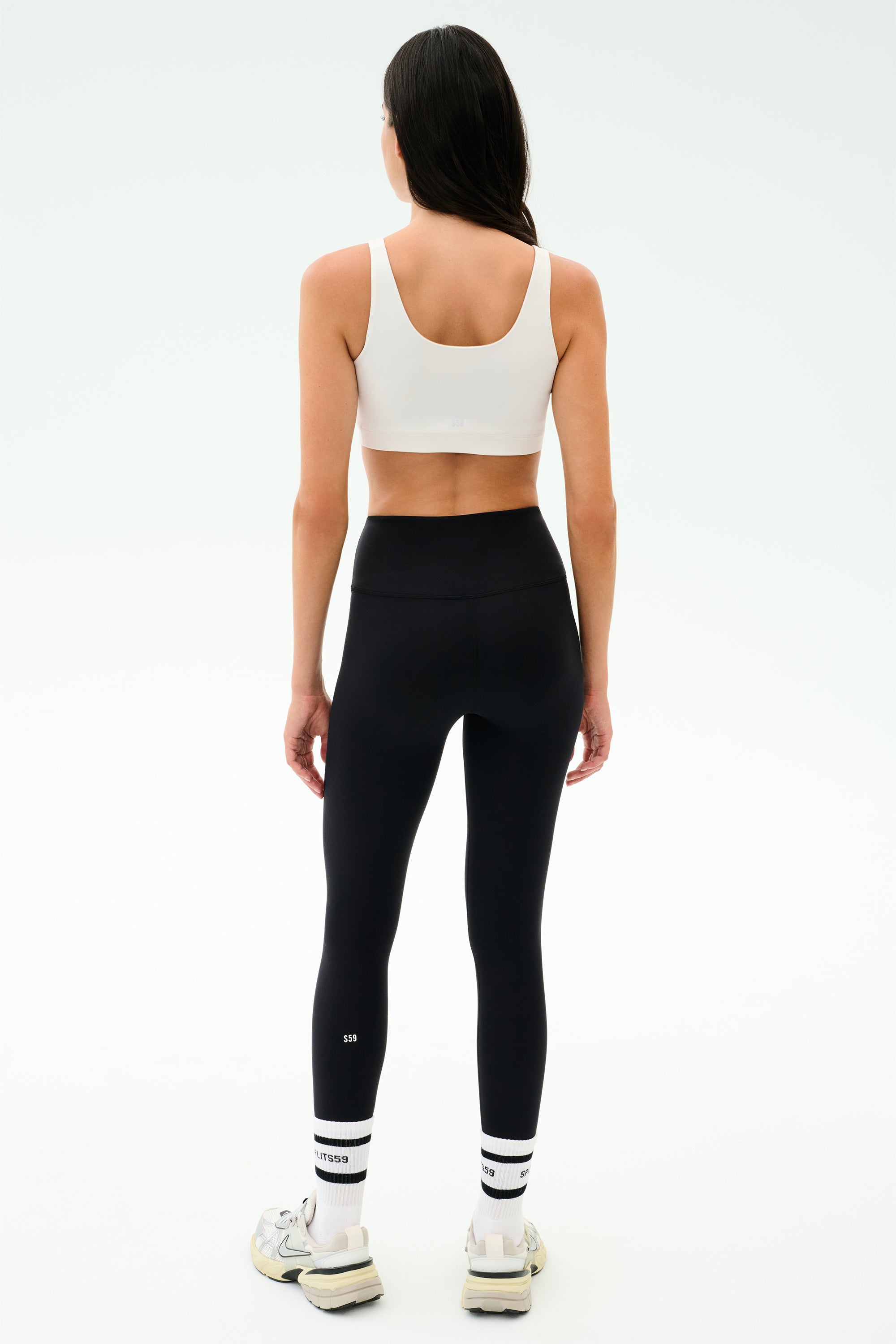 A woman, seen from behind, wears the SPLITS59 Sprint Rigor Bra in White, black high-waisted leggings, white socks, and sneakers—poised for a workout against a plain light background.