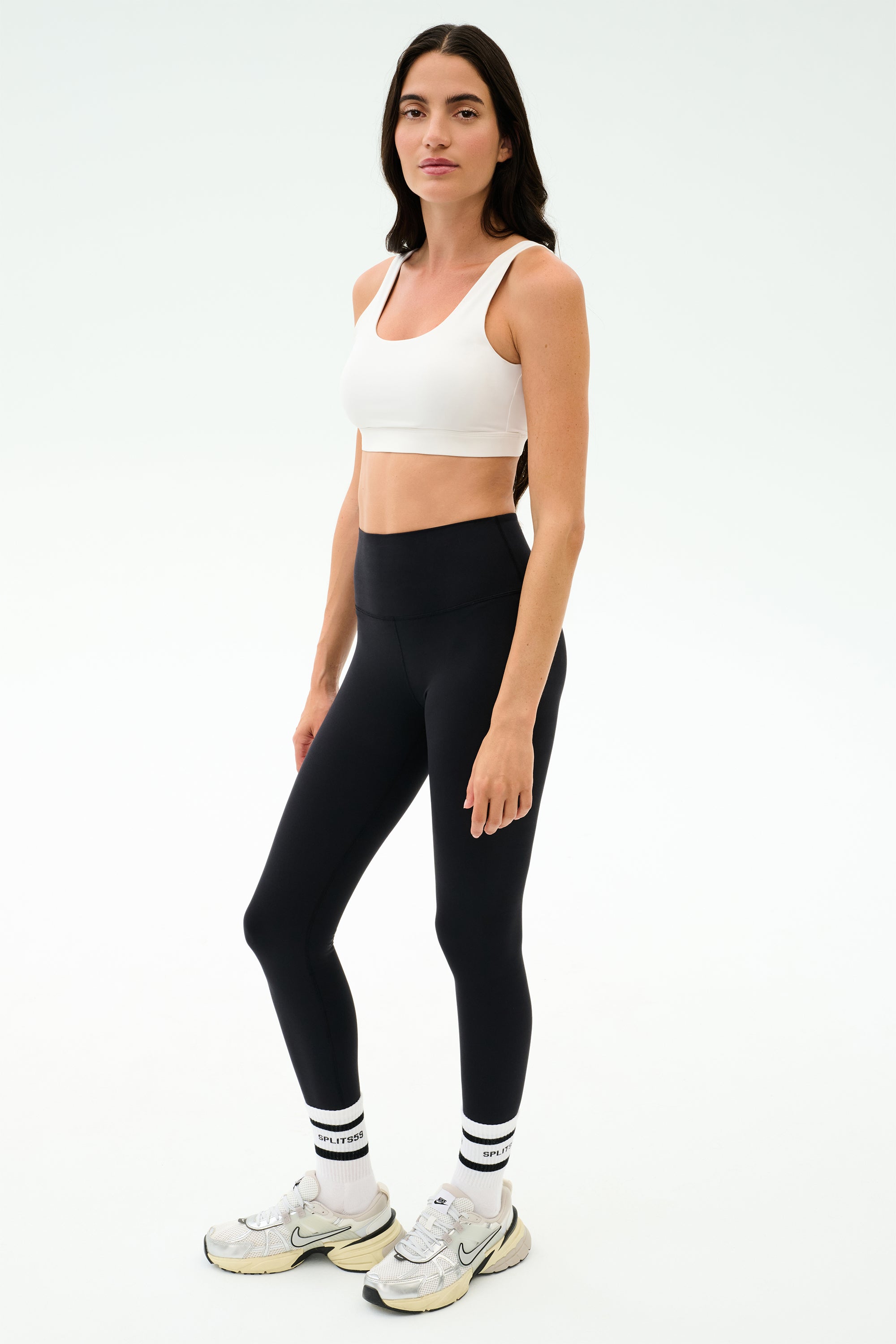 A woman poses against a plain backdrop in the SPLITS59 Sprint Rigor Bra - White—perfect for gym sessions—styled with black leggings, white socks featuring black stripes, and white sneakers.