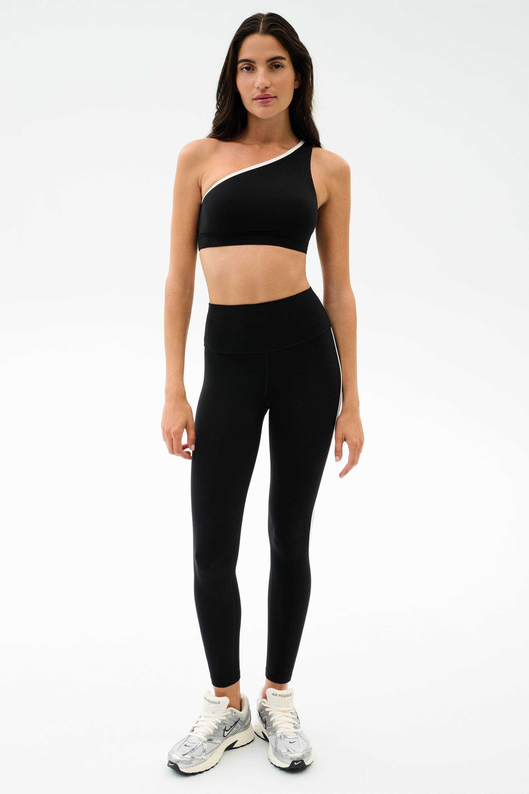 A woman stands in a studio wearing the SPLITS59 Josie Airweight One Shoulder Bra in Black/White with matching high-waisted leggings and gray athletic shoes—ideal for hot yoga—against a plain white background.