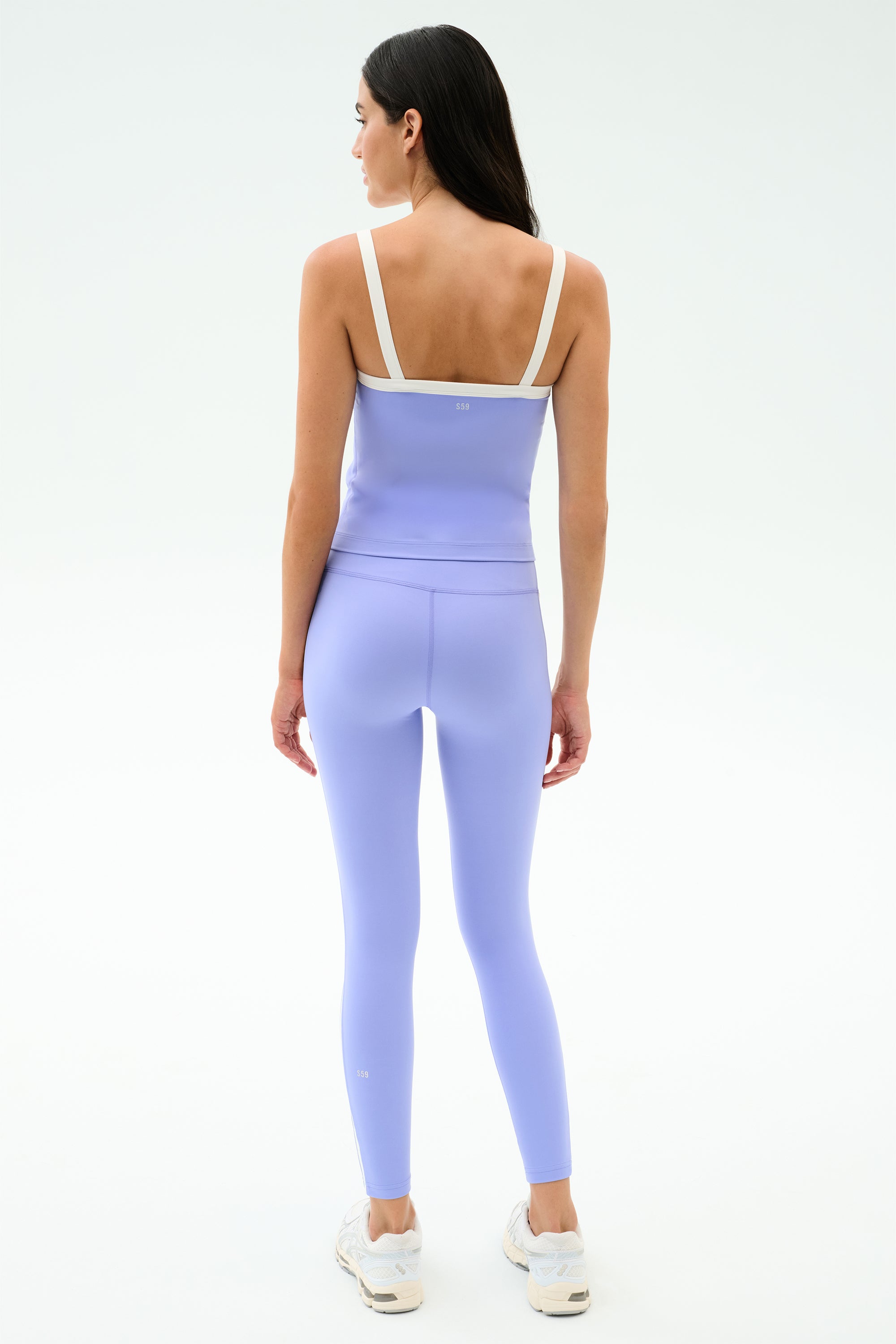 A woman faces away on a white background, wearing SPLITS59's Clare High Waist Rigor 7/8 leggings in Purple Haze/White—ideal for the gym—paired with a light purple athletic top and white sneakers.
