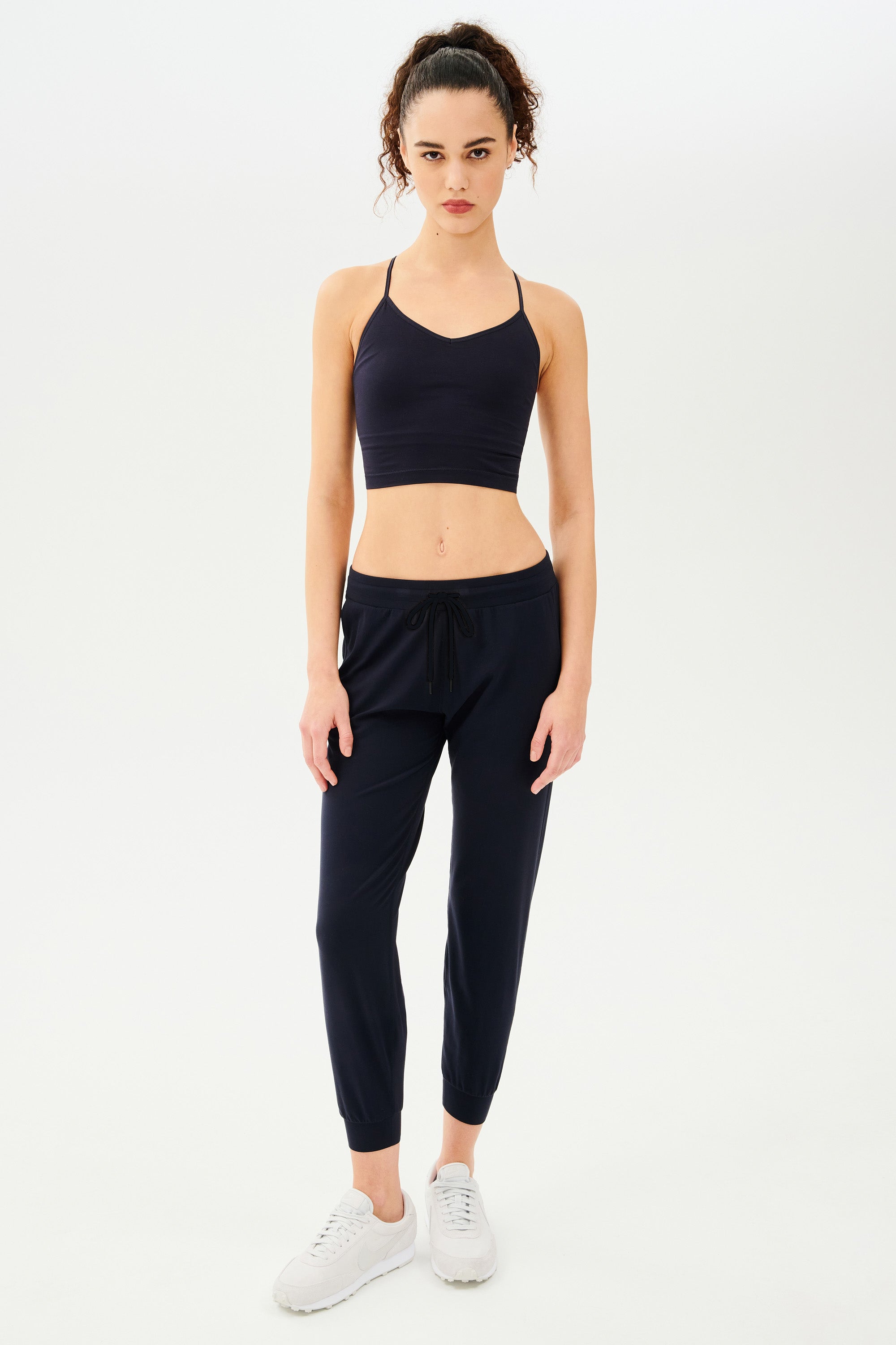 Full front view of girl wearing dark blue sweatpants with black tie around waistband and a dark blue sports bra with white shoes