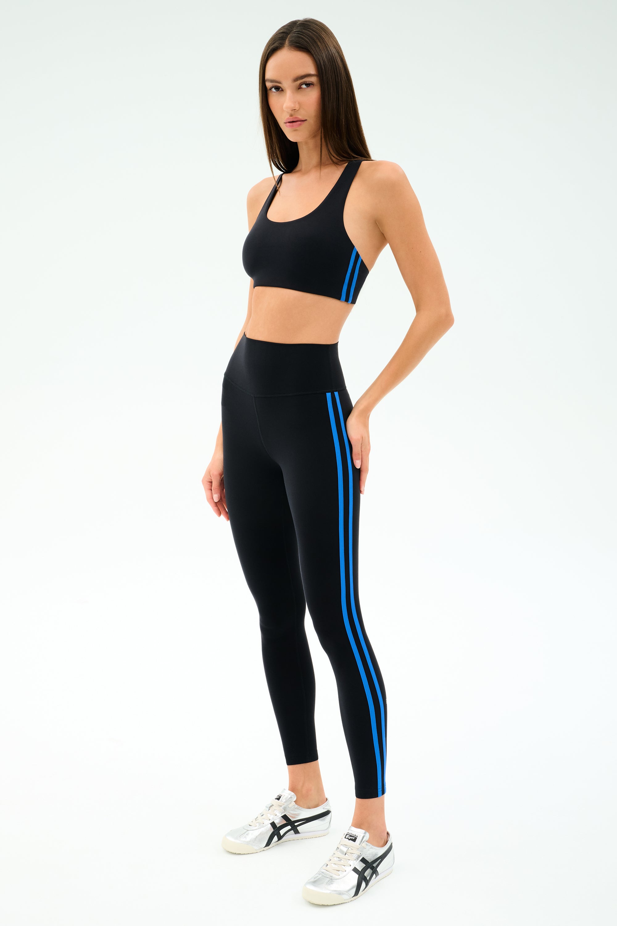 A woman models the SPLITS59 Ella High Waist Airweight 7/8 Black/Classic Blue leggings with a matching black sports bra and white sneakers, standing against a plain white background.