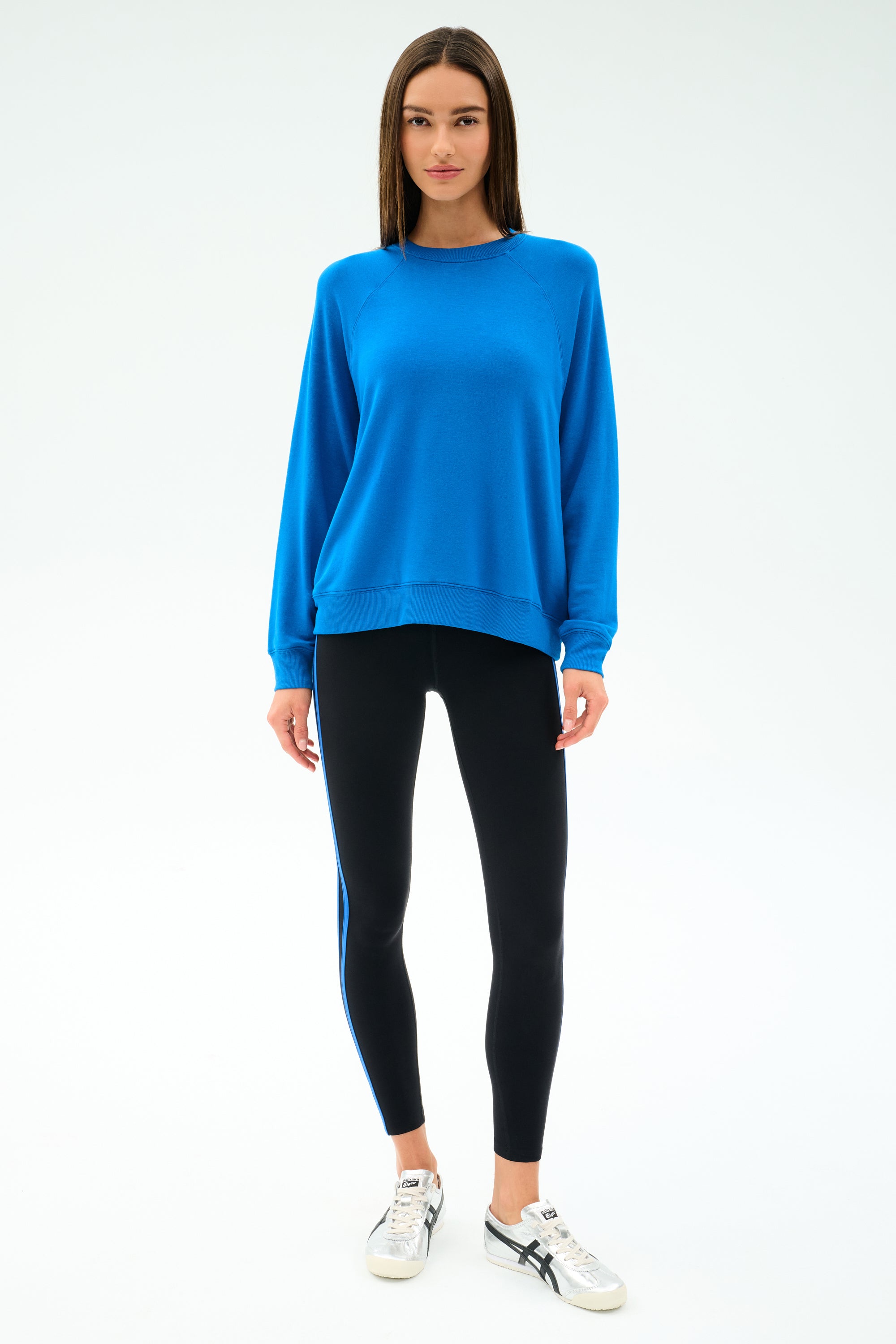 A woman faces forward against a plain light background, wearing the SPLITS59 Andie Oversize Fleece Sweatshirt in Classic Blue and black leggings with a blue stripe, paired with white sneakers.