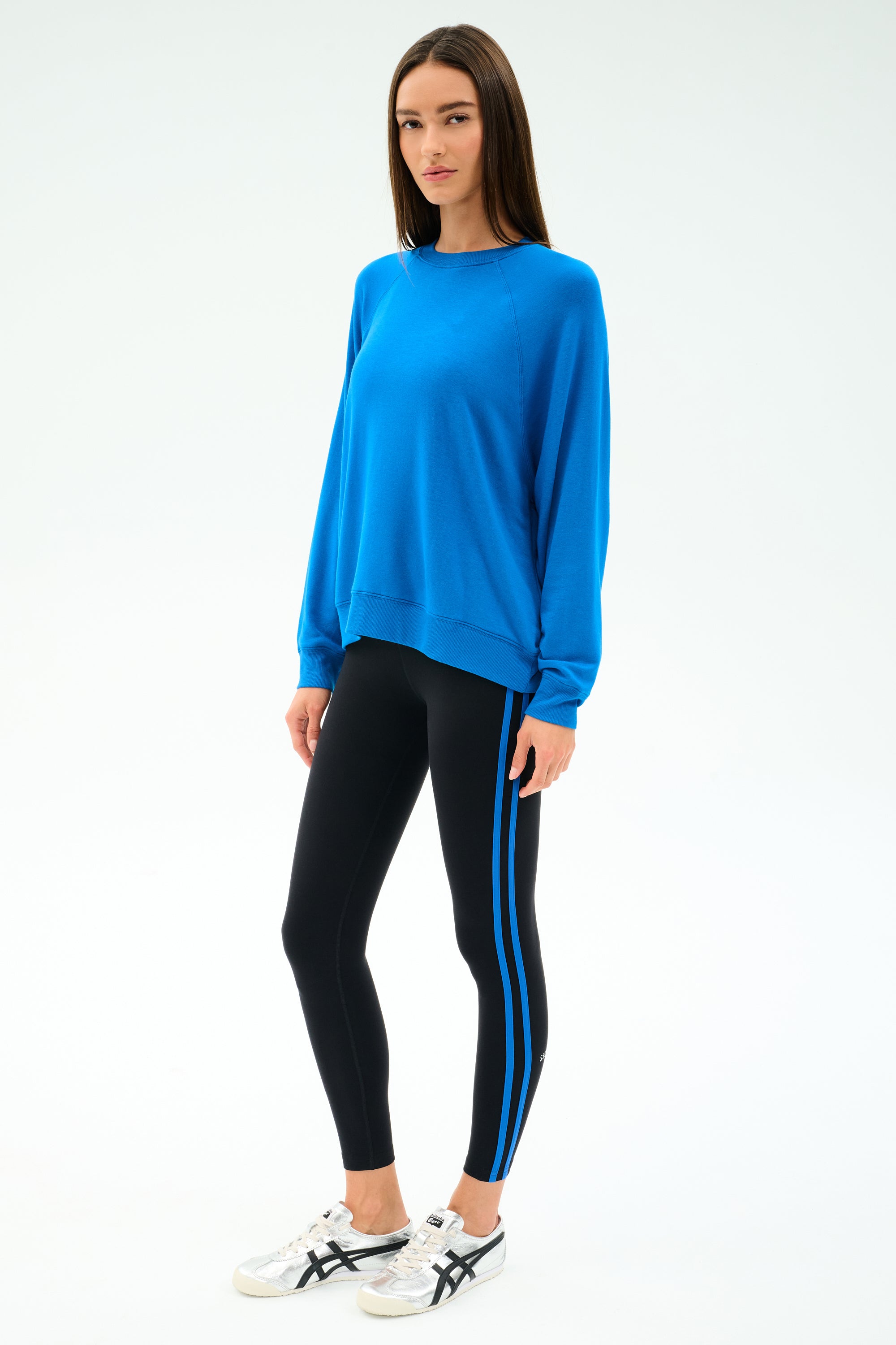 A person wears the SPLITS59 Andie Oversize Fleece Sweatshirt in Classic Blue and black leggings with blue stripes, made from a soft modal spandex blend, paired with white sneakers against a plain white background.