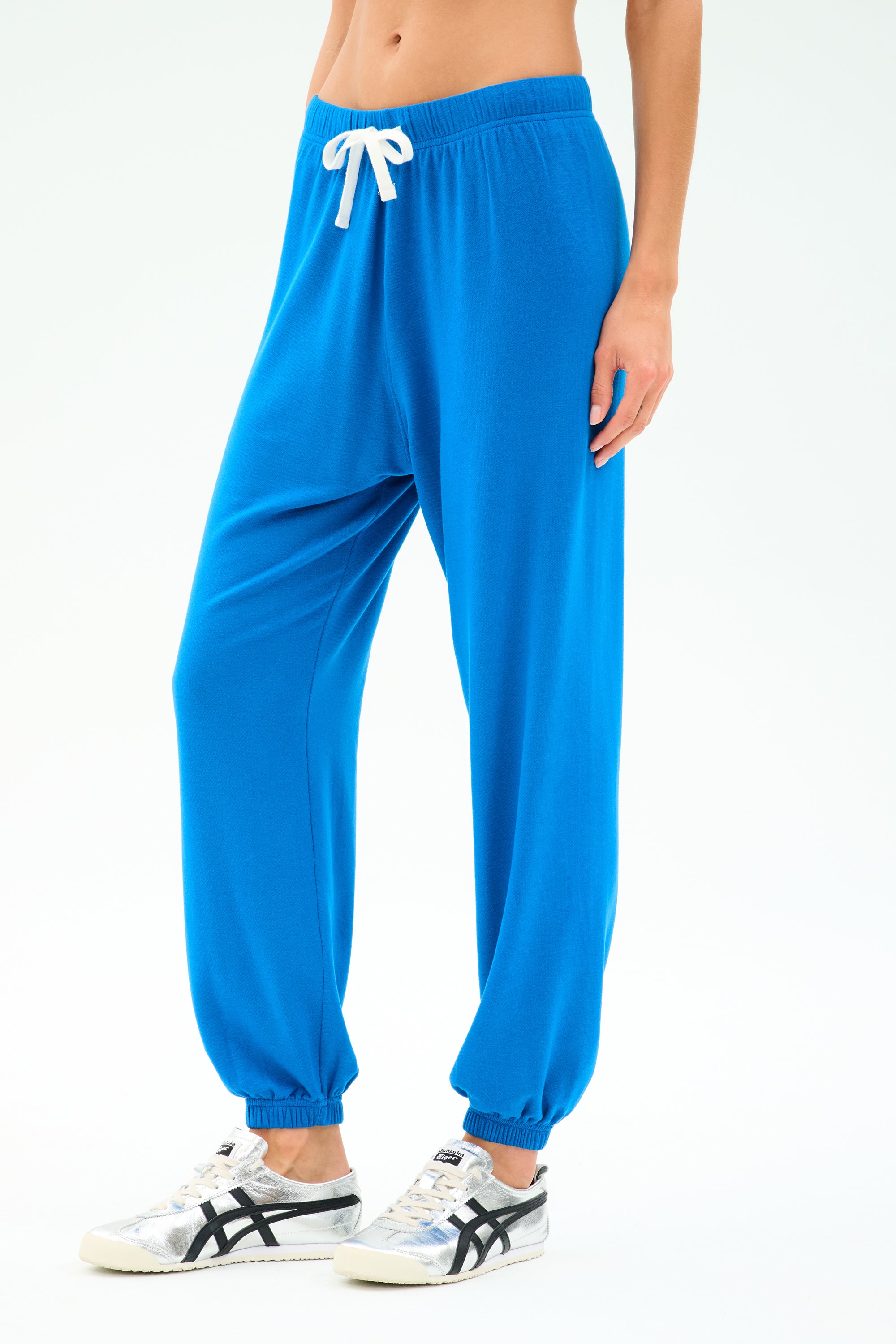 Person wearing SPLITS59 Andie Oversized Fleece Sweatpant in Classic Blue with a white drawstring and white sneakers with black details, standing against a plain light background.