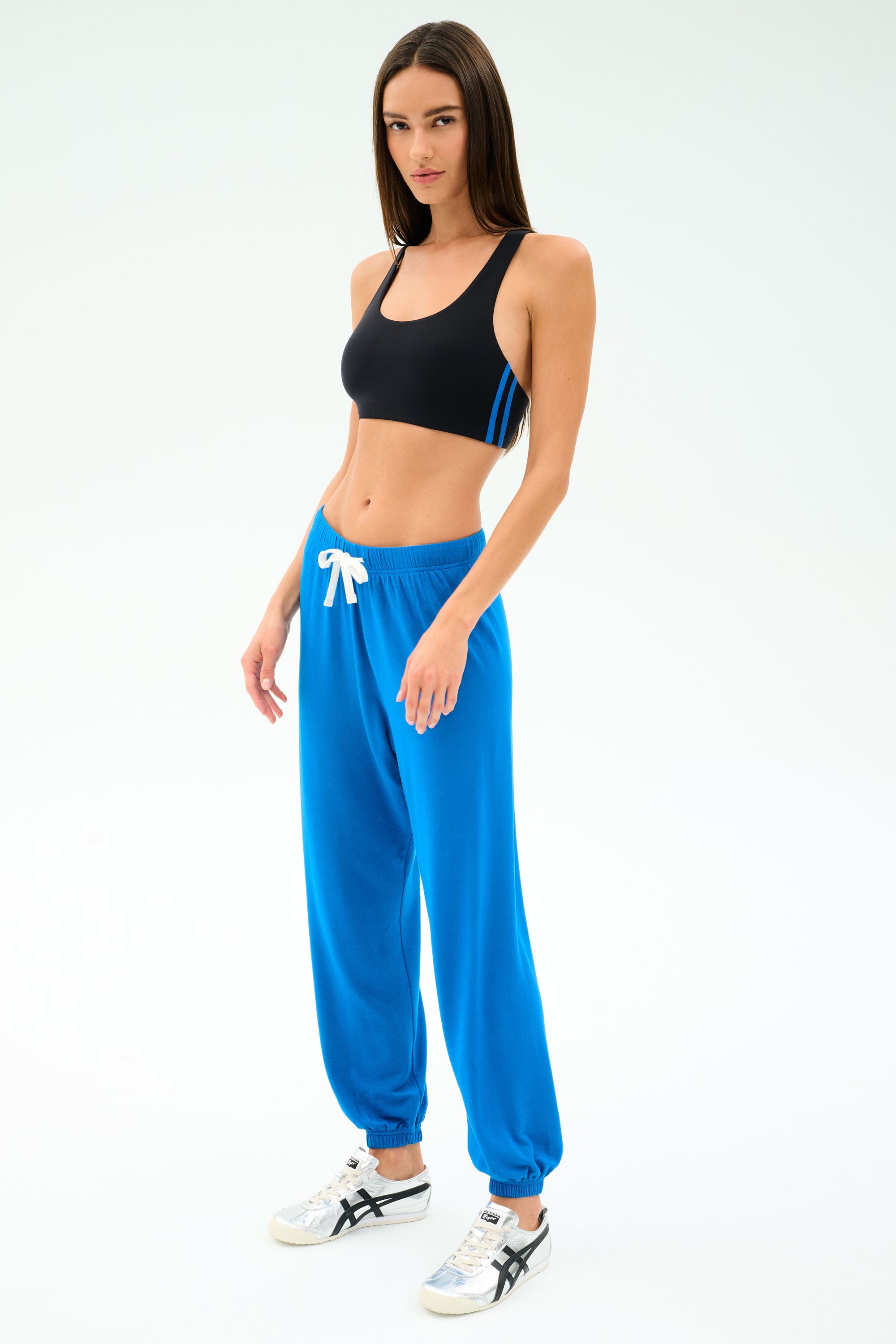 A woman in a black spandex sports bra, SPLITS59 Andie Oversized Fleece Sweatpant in Classic Blue with a white drawstring, and white sneakers stands against a plain light background.