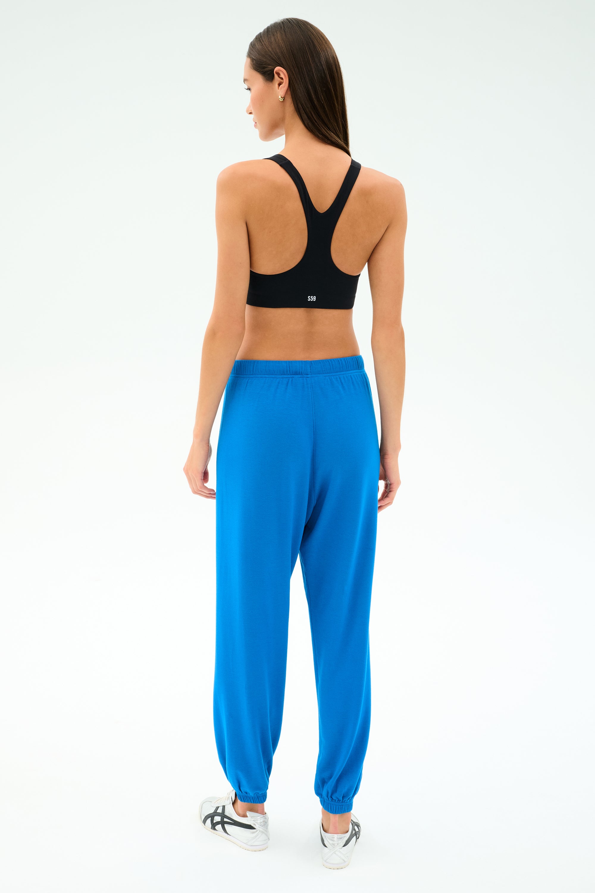 A person stands facing away, wearing a black spandex sports bra, white sneakers, and SPLITS59's Andie Oversized Fleece Sweatpant in Classic Blue against a plain light background.