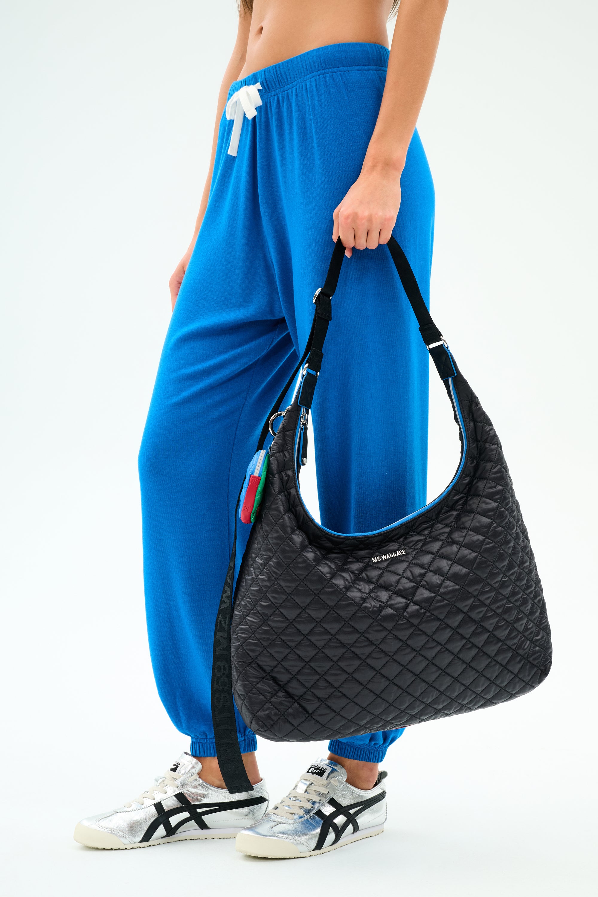A person in blue jogger pants and white sneakers carries the SPLITS59 MZW x Splits59 Carry All, a large black quilted shoulder bag with a strap and small decorative keychains.