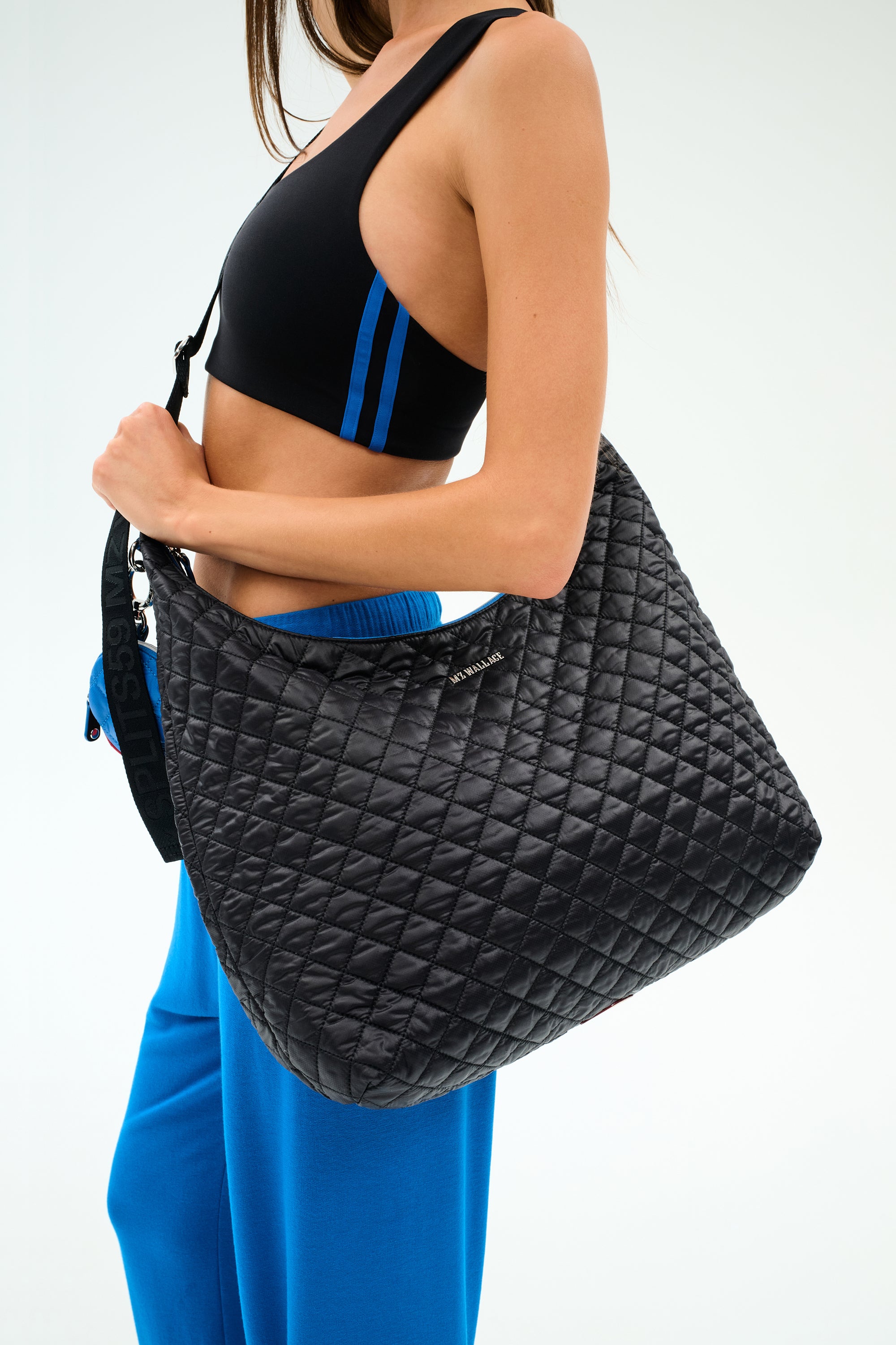 A woman in a black sports bra and blue pants carries the SPLITS59 MZW x Splits59 Carry All, a large black quilted shoulder bag with a wide strap.