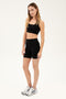 Full side front view of woman with dark blonde straight hair wearing black bra and black high waist bike shorts paired with white shoes with black stripes