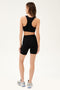Full back view of woman with dark blonde straight hair wearing black bra with racer straps and black high waist bike shorts paired with white shoes with black stripes