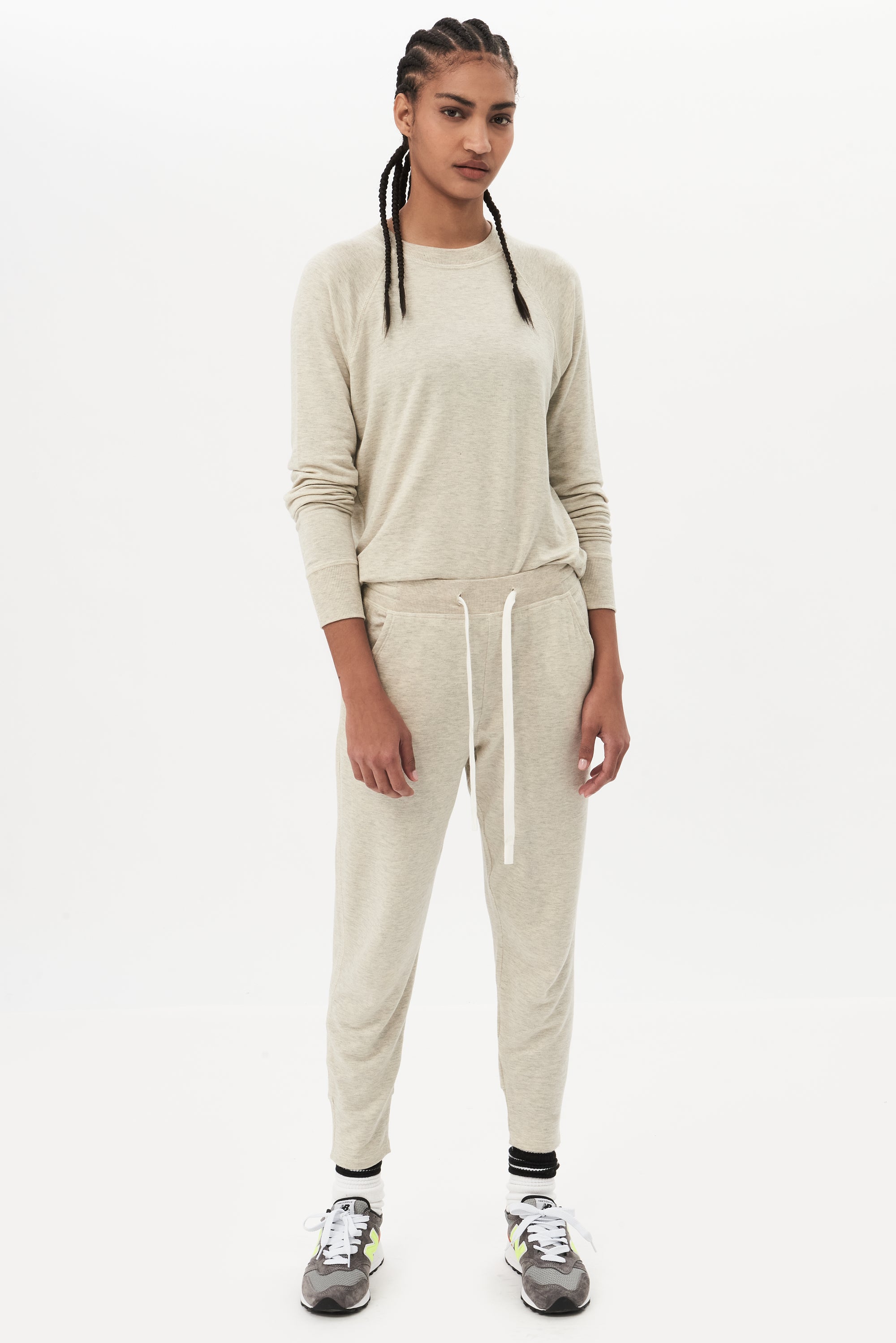 Full front view of woman with black braided hair hair wearing a light gray sweatpant with tapered leg and above ankle length with white drawstring and side hip pockets and light grey crewneck sweatshirt. Paired with dark gray shoes with a bright green and red color block.