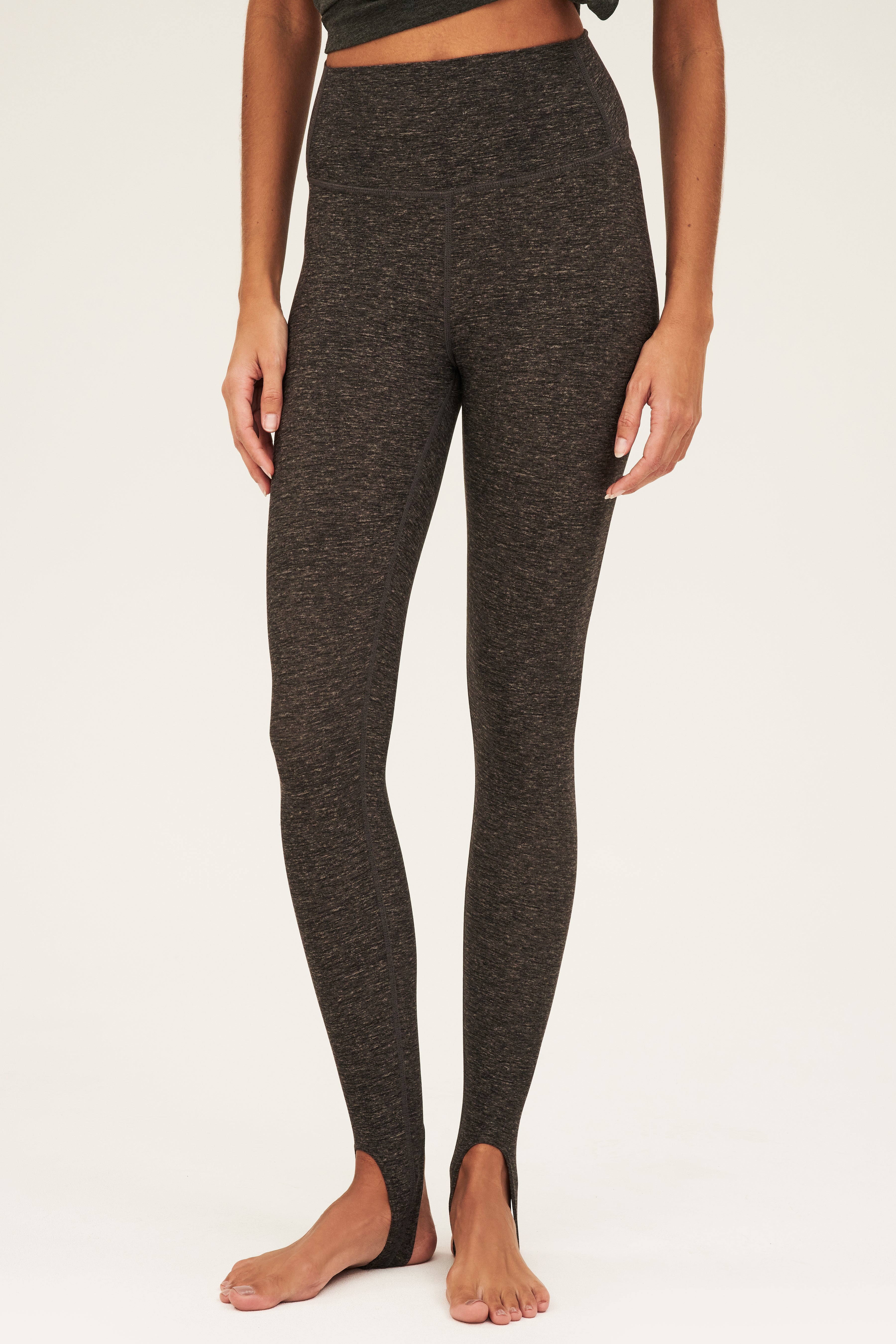 Front view of woman wearing dark grey jersey tank with side tie and dark gray and white speck leggings with bottom strap that wraps around the foot on model's bare feet